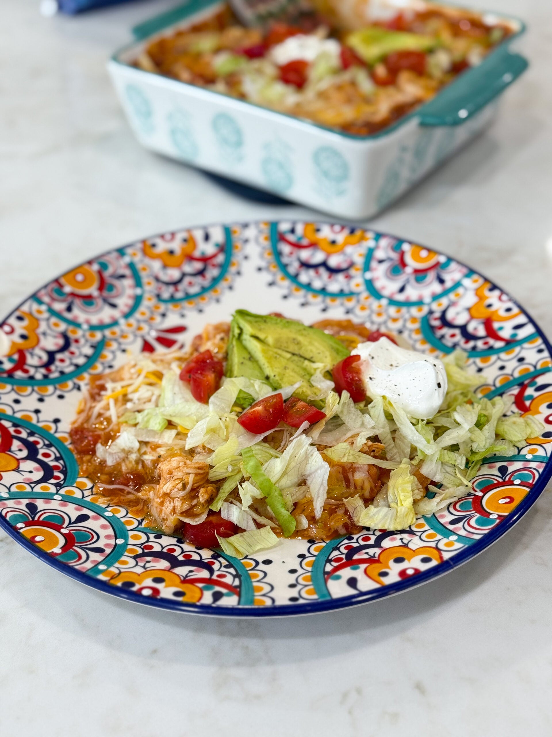 Baked red chicken enchiladas in a on a plate topped with tomato, sour cream, lettuce and avacado.