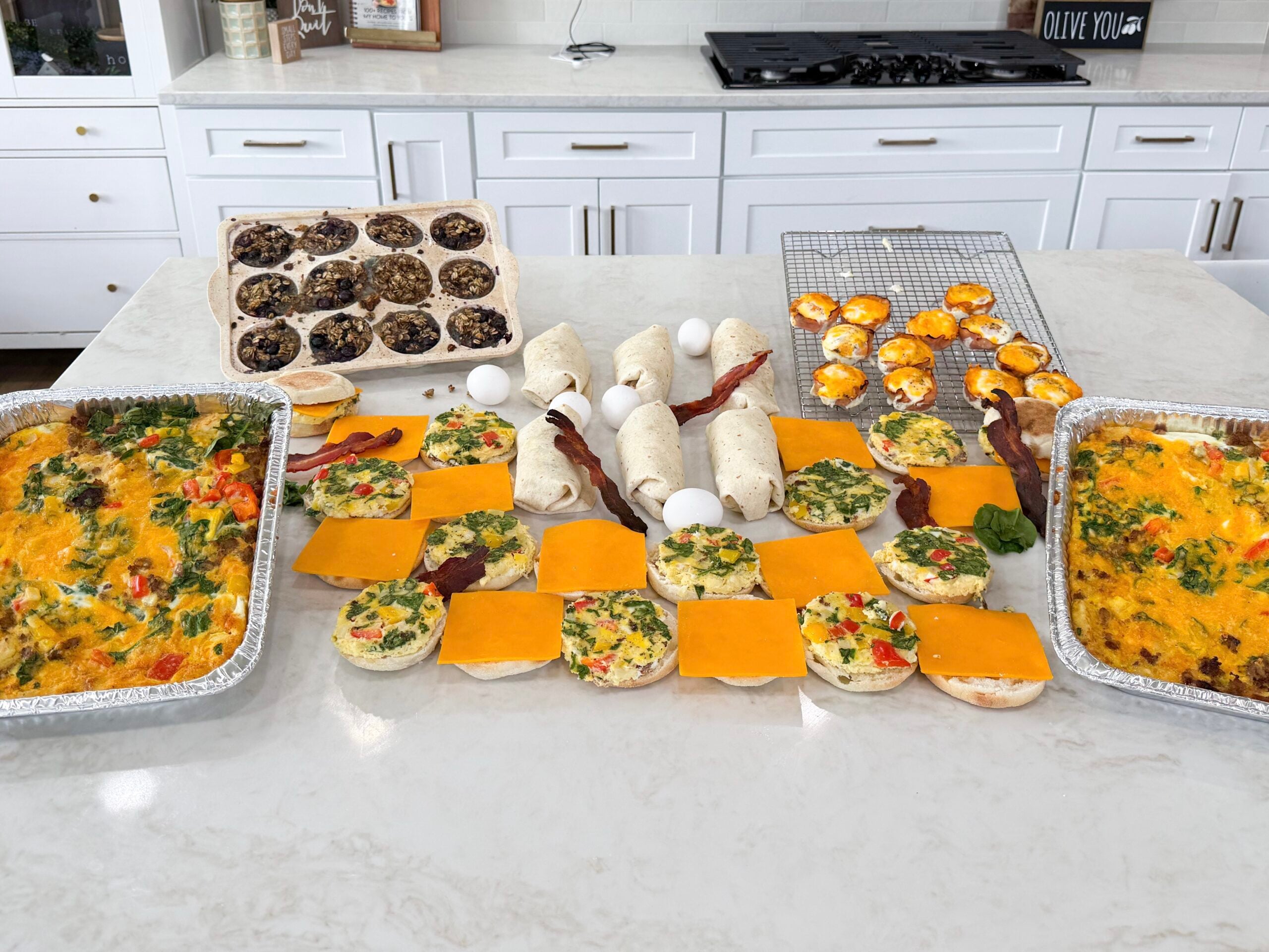 Completed breakfast meal prep including two breakfast casseroles, baked blueberry oatmeal cups in a muffin tin, ham and egg bites on a cooling rack, breakfast burritos, and breakfast sandwiches on a white countertop.