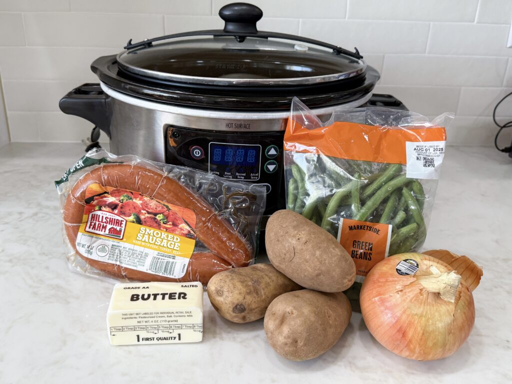 large black and grey slow cooker with sausage, green beans, onion, butter, and potatoes on a white counter