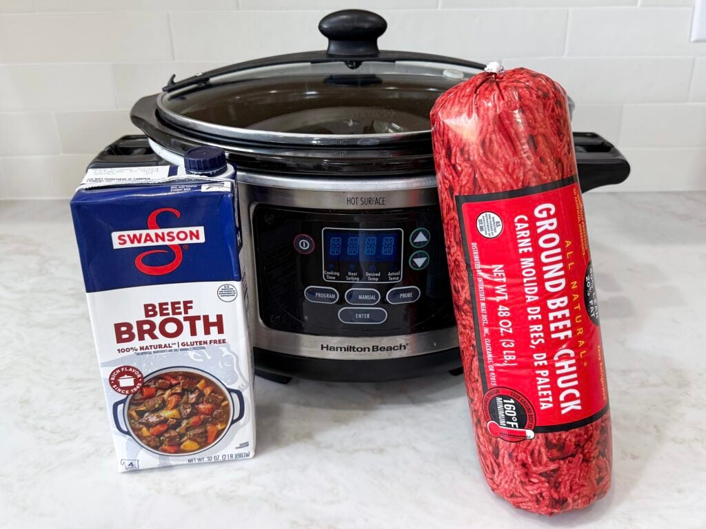 large slow cooker on a white counter with beef broth and package of ground beef