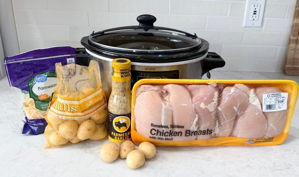 Black and grey slow cooker on a counter with raw chicken, potatoes, with buffalo wing wings sauce and parmesan cheese